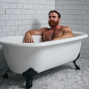 A bodybuilder man is sitting in a old style bathtub that is whit...