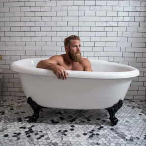 A bodybuilder man is sitting in an old-style bathtub that is whi...