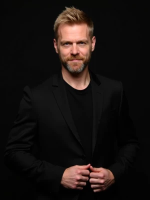 A man with short blonde hair and a beard, dressed in a black sui...