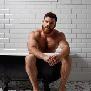 A bodybuilder man is sitting in a old style bathtub that is whit...